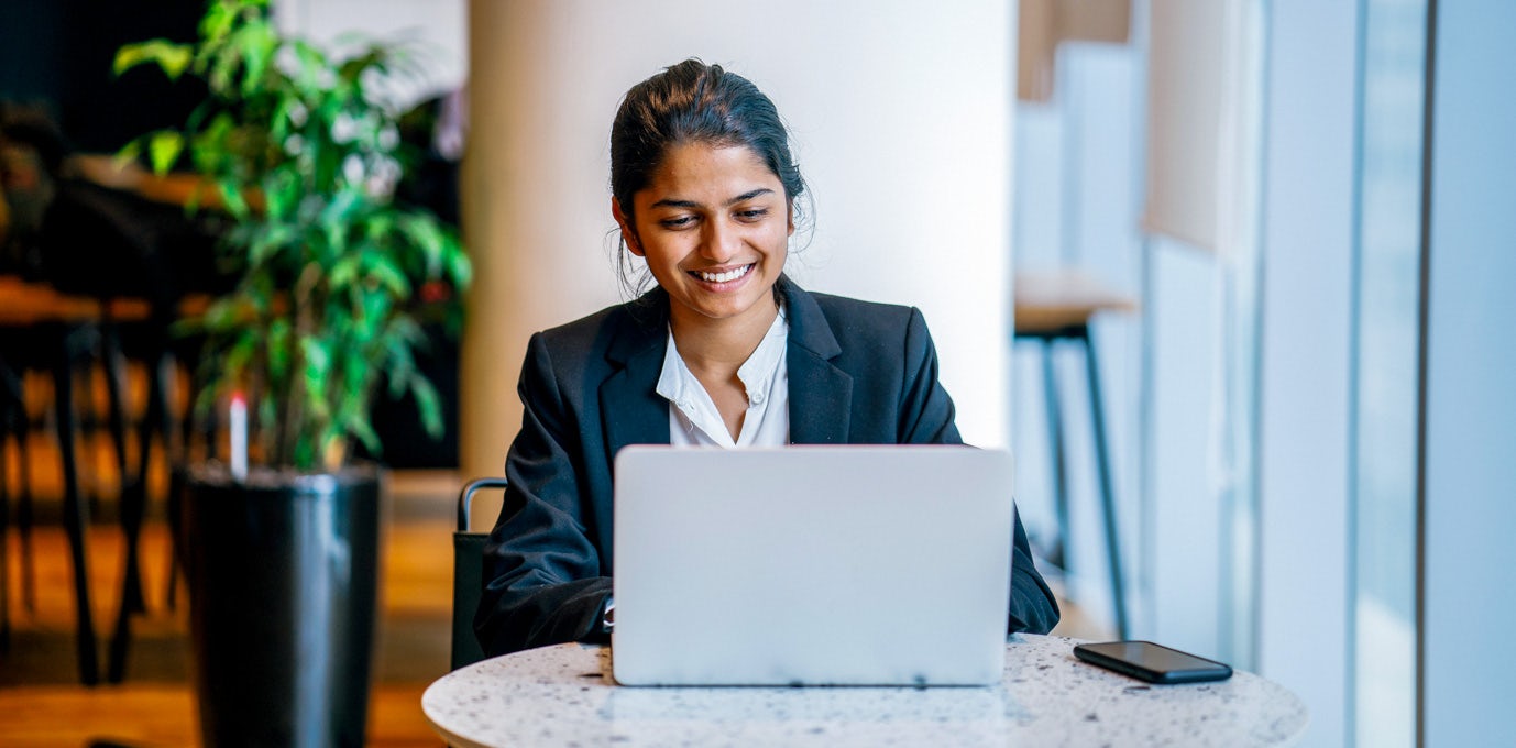 [Imagen destacada] Mujer sonriente con traje de negocios sentada en una mesa con su laptop y smartphone.