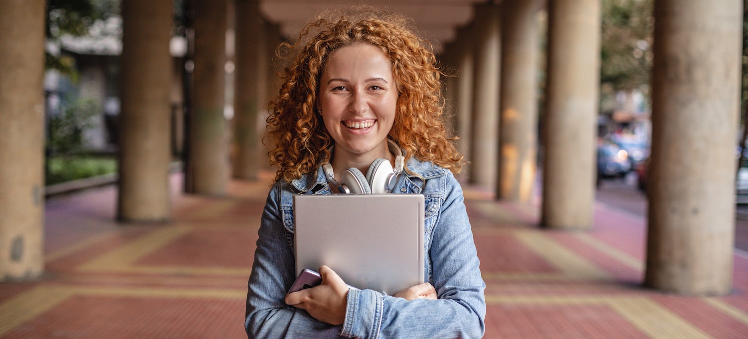[Featured image] A young white woman with curly orange hair stands outside in a portico holding a notebook and smiling. 