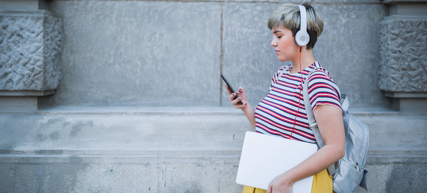 [Featured image] A young woman wearing a striped shirt and listening to over-the-ear headphones walks while using her cell phone. 