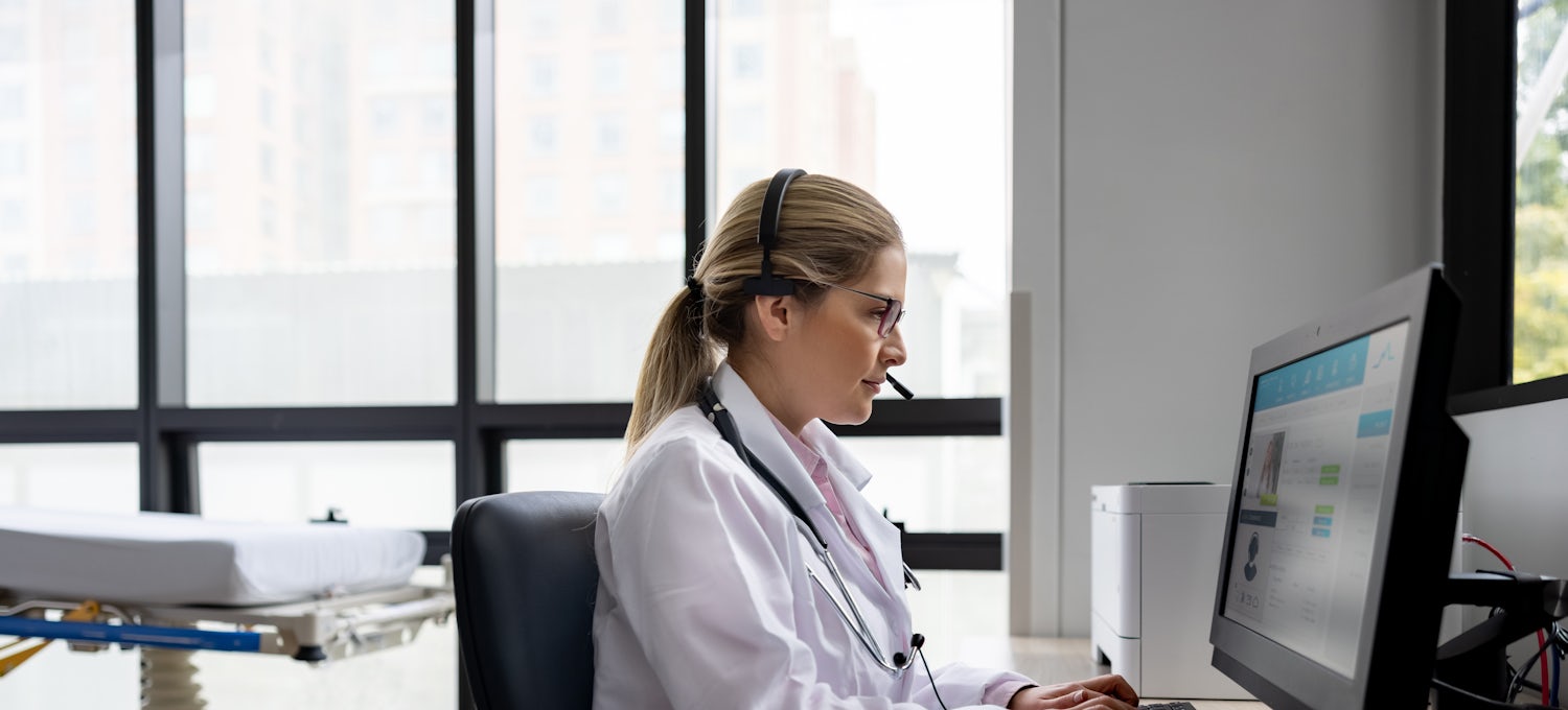 [Featured Image]:  A female telehealth nurse, wearing a white uniform, is sitting in front of her computer, communicating with a patient.