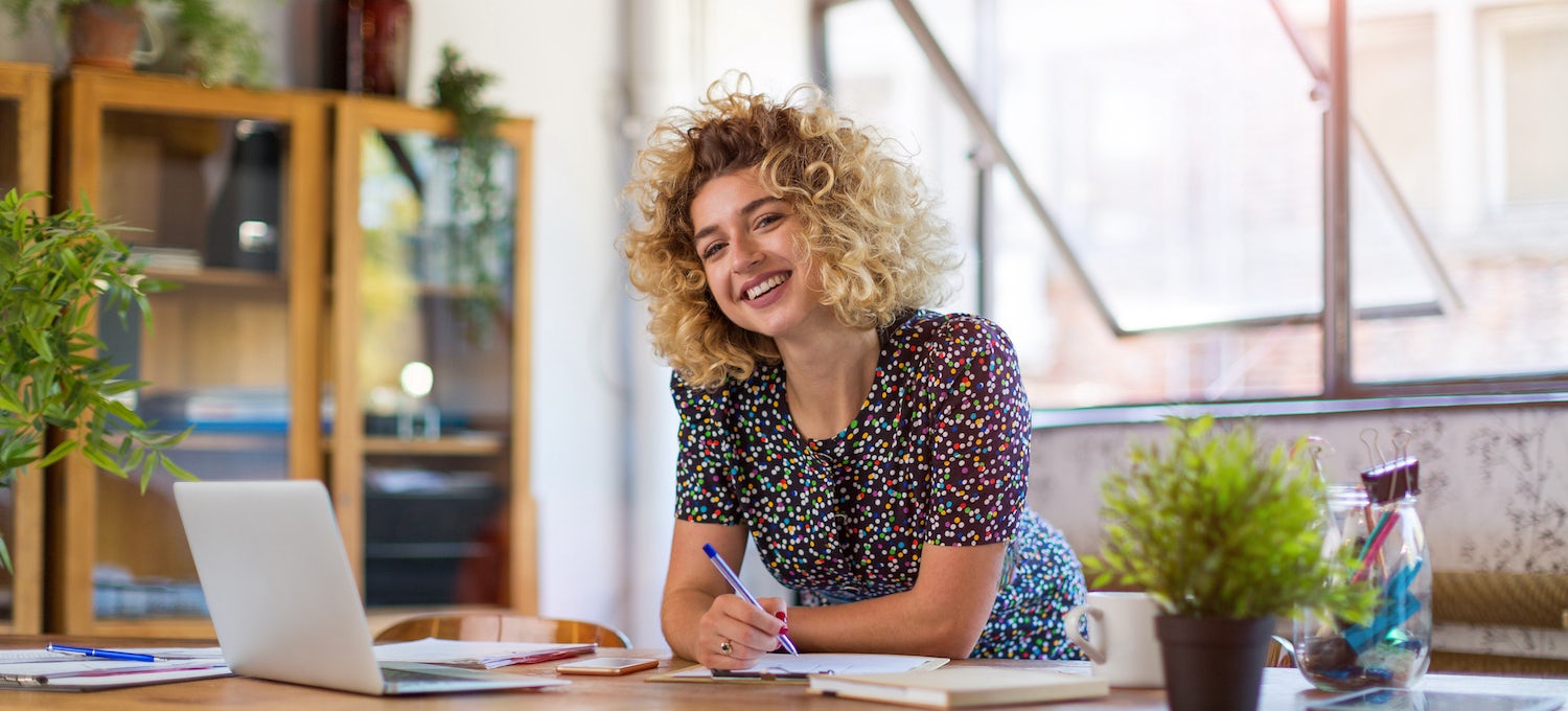 [Featured image] A young woman with curly hair leans over a table with a laptop on it, holding a pen, and smiles into the camera. 