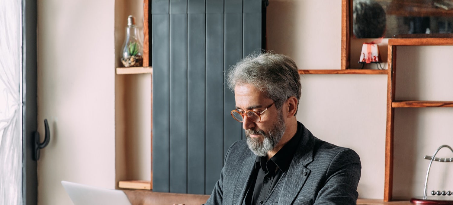 [Featured image] A man with grey hair and a beard wearing a grey sports jacket, black shirt, and glasses is sitting at a desk working on a laptop. 