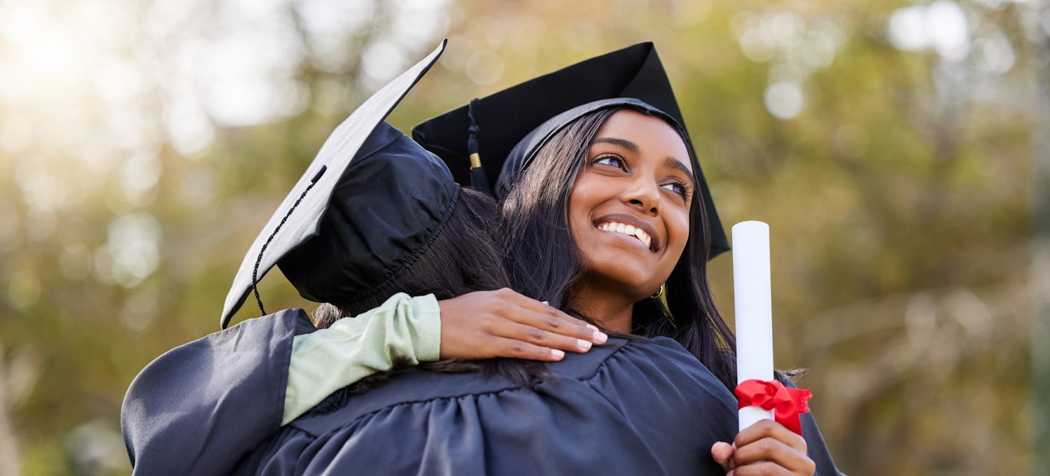 [Imagen destacada] Dos graduadas con toga y birrete negros y una sosteniendo un diploma celebran la obtención de sus títulos de maestría.