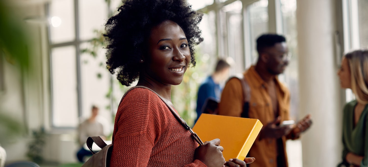 [Featured image] A Black female doctorate student in a coral pink sweater smiles into the camera while holding a yellow folder. 