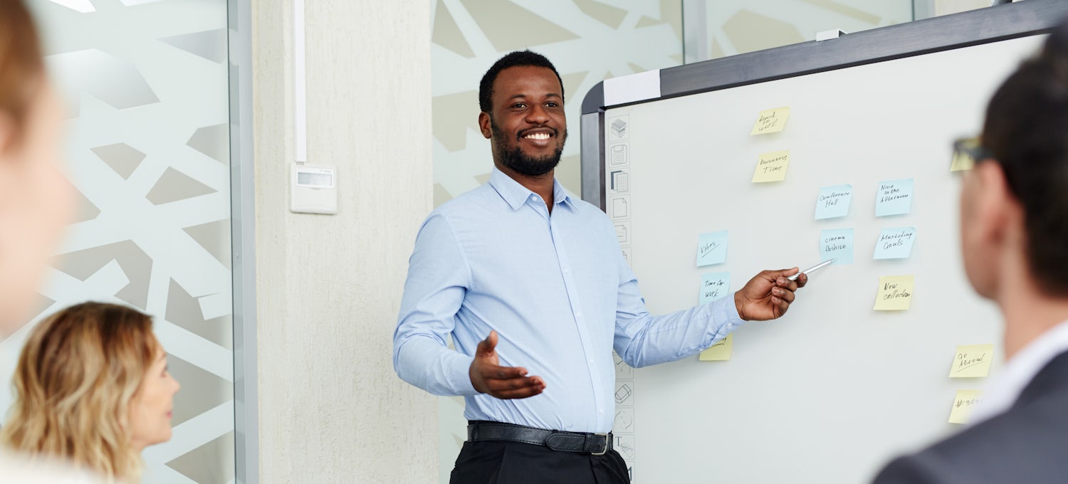 [Featured image] A project manager in blue button-down shirt gestures toward a whiteboard covered in colorful sticky notes while presenting to a team.