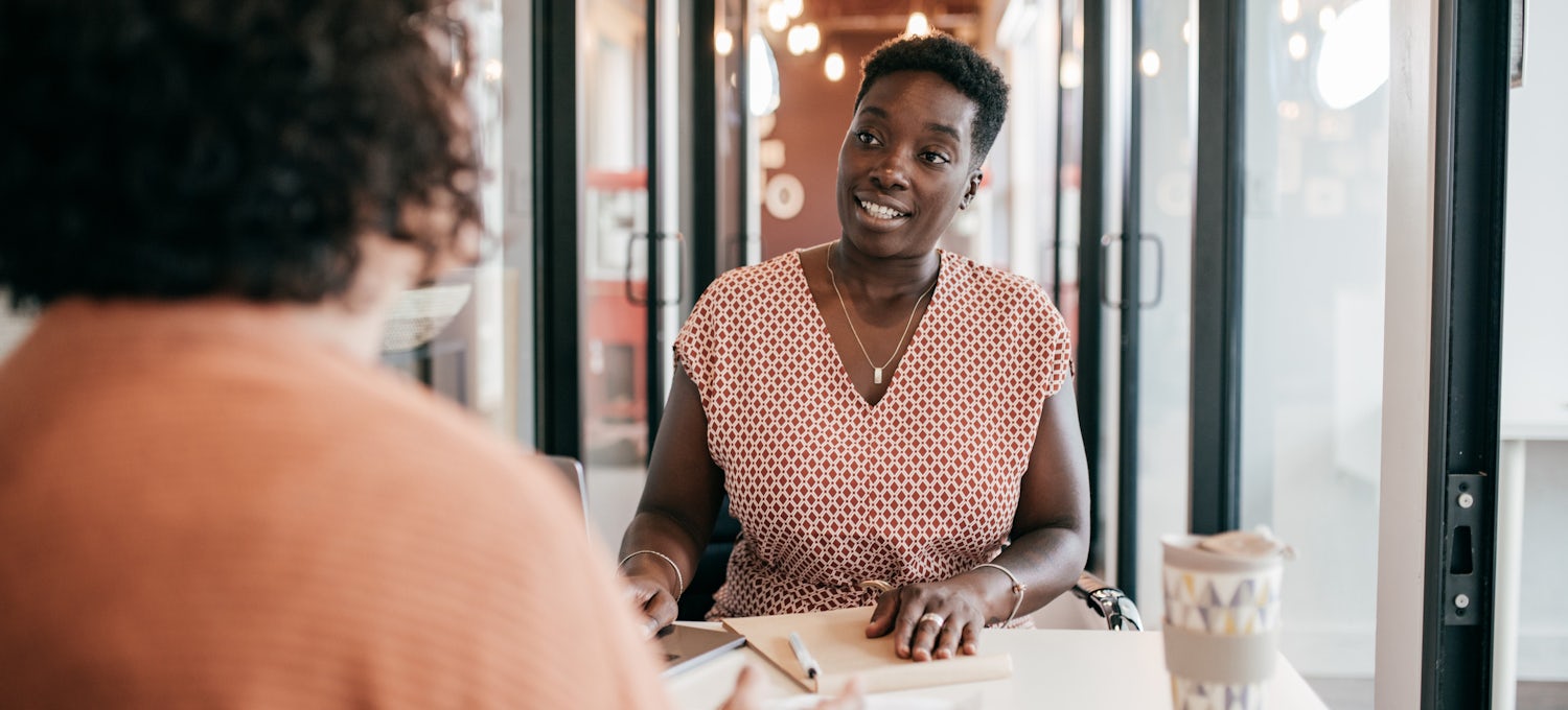 [featured image] A black woman wearing a pink patterned dress sits across the table from another woman in an office.