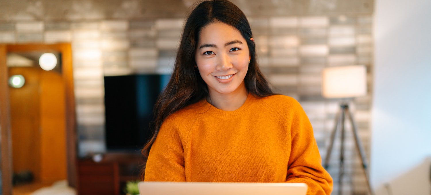 [Featured image] A young woman in a burnt-yellow sweater smiles into the camera while holding her laptop. 