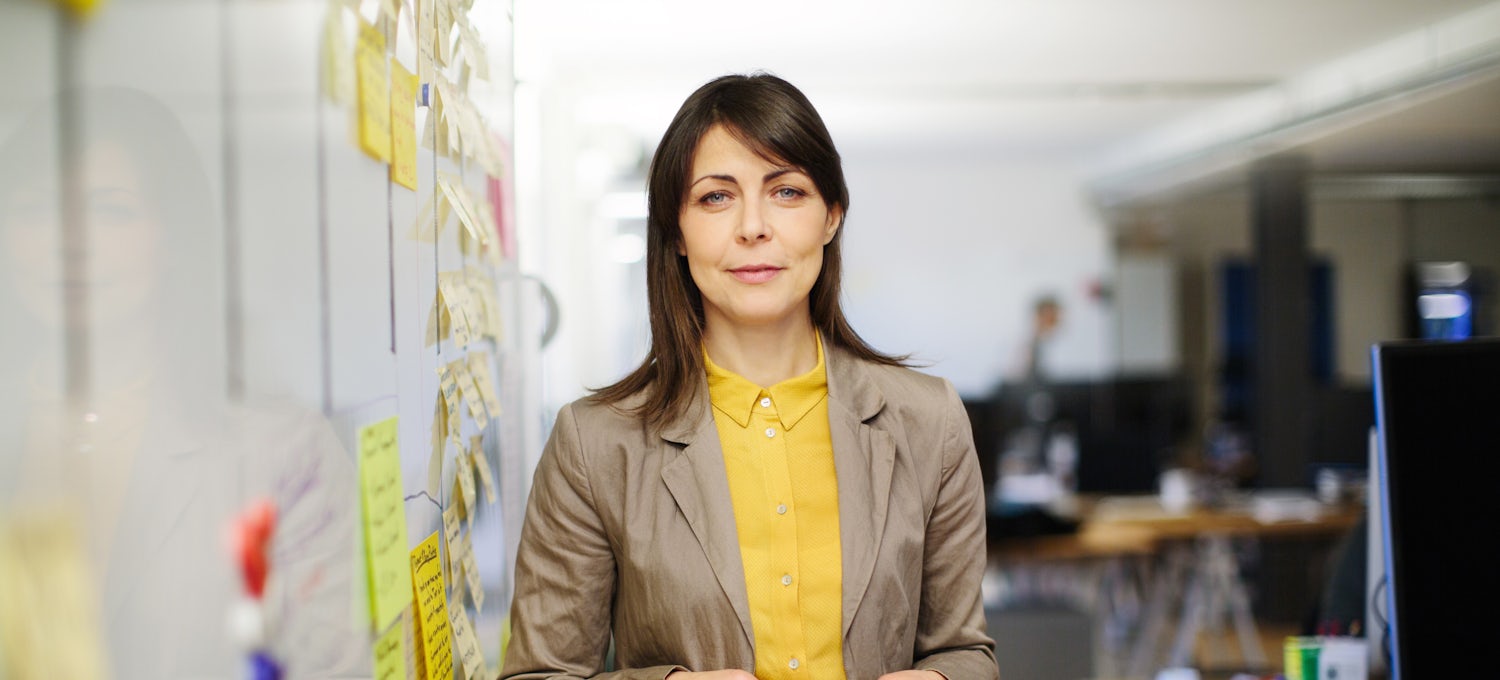 [Featured Image] An Agile coach wears a brown blazer and yellow blouse and stands next to a whiteboard with post-it notes on it.