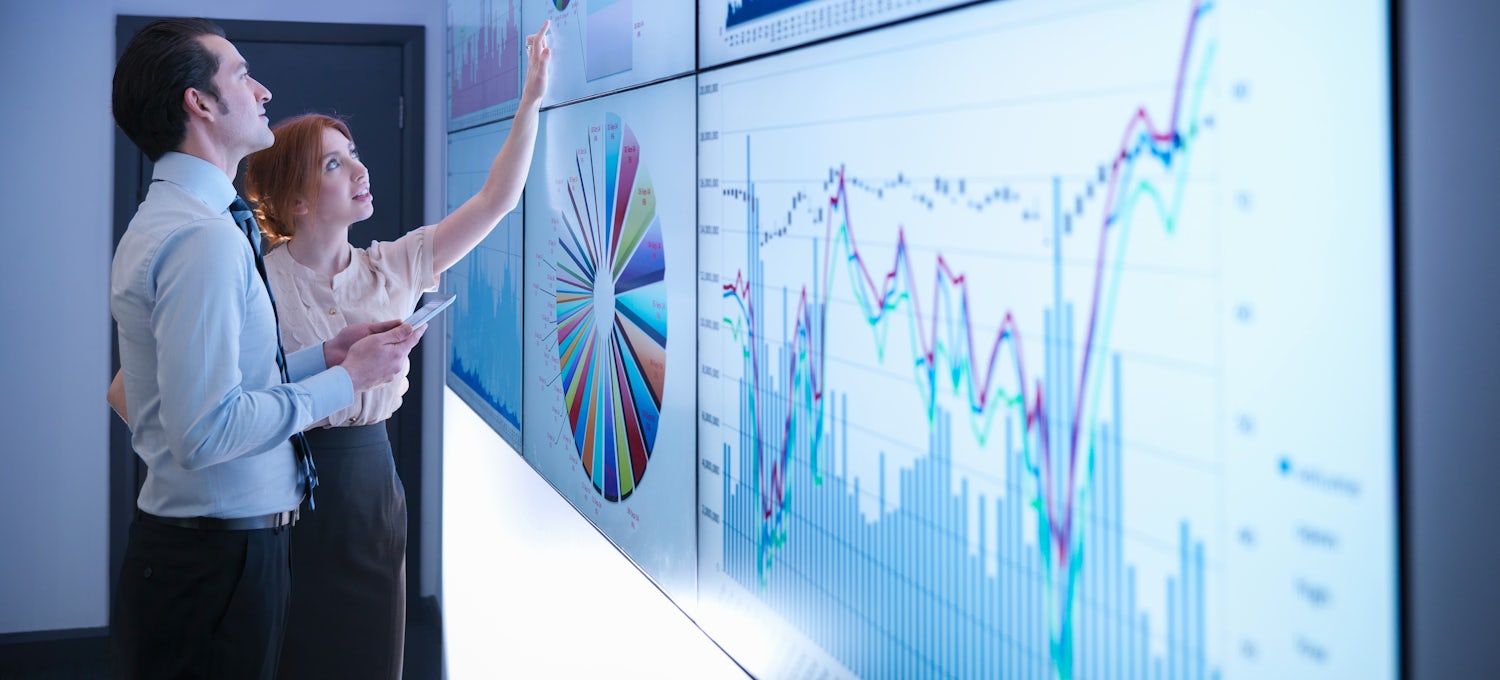 [Featured Image] Two dala analysts examine charts and graphs projected on a white smart board.