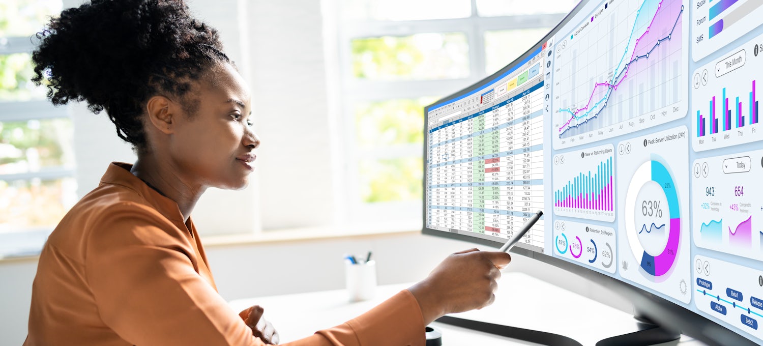 [Featured Image] Woman in a rust-colored long-sleeved shirt looking at computer screens displaying business intelligence data for risk assessment.
