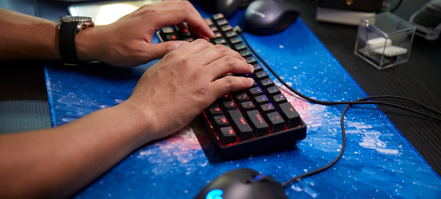 [Featured Image] Two hands hover over a colorful light-up keyboard sitting on a galaxy-printed desk mat next to a computer mouse.