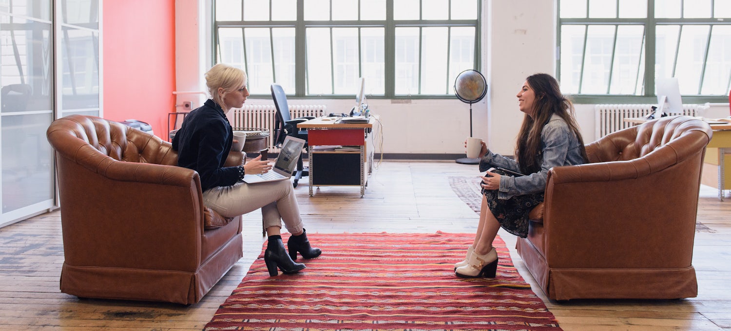 [Featured Image] Two women have a conversation in an office. 