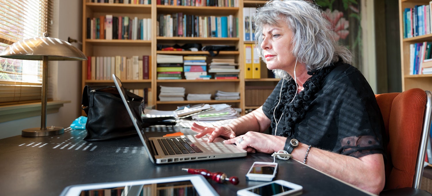 [Featured Image] A woman has earbuds on and is using her laptop at her home library.  