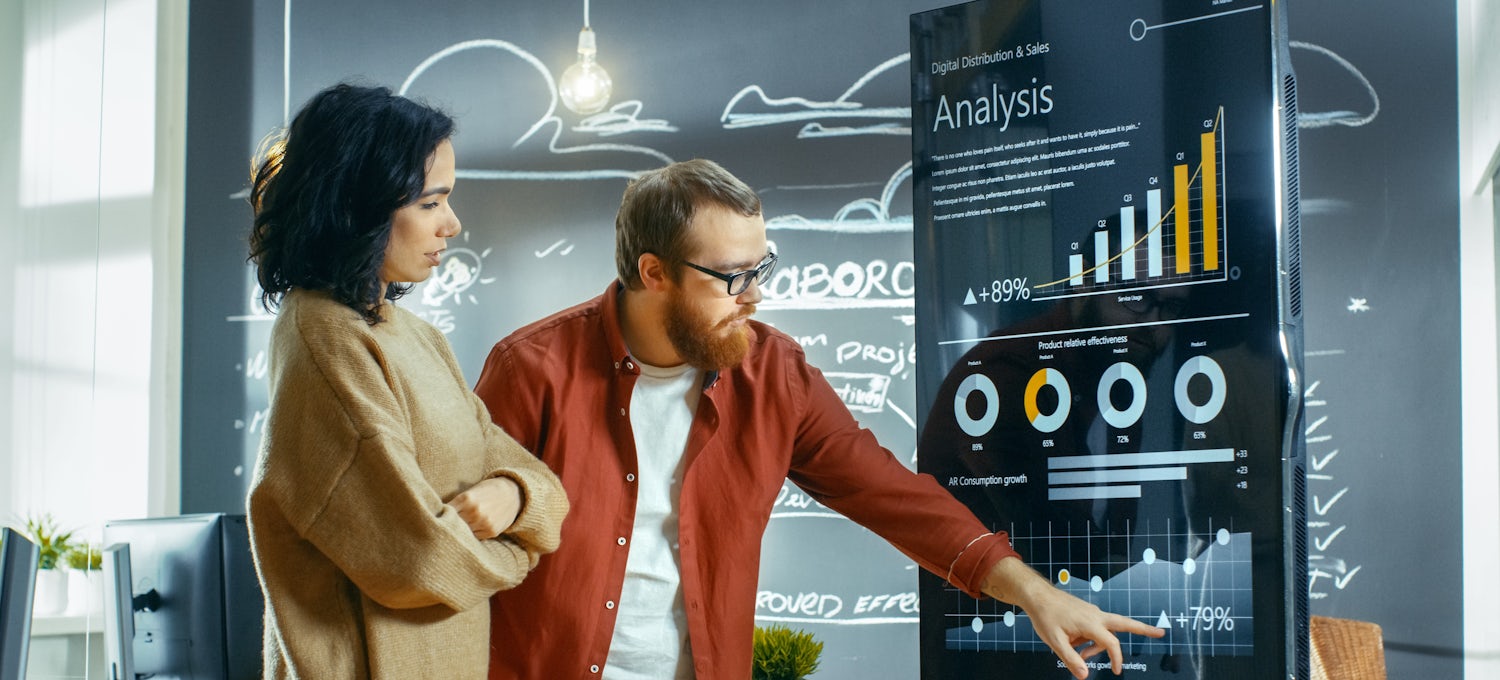 [Featured Image] Two coworkers review sales forecasting data on a large screen display in an open workspace.
