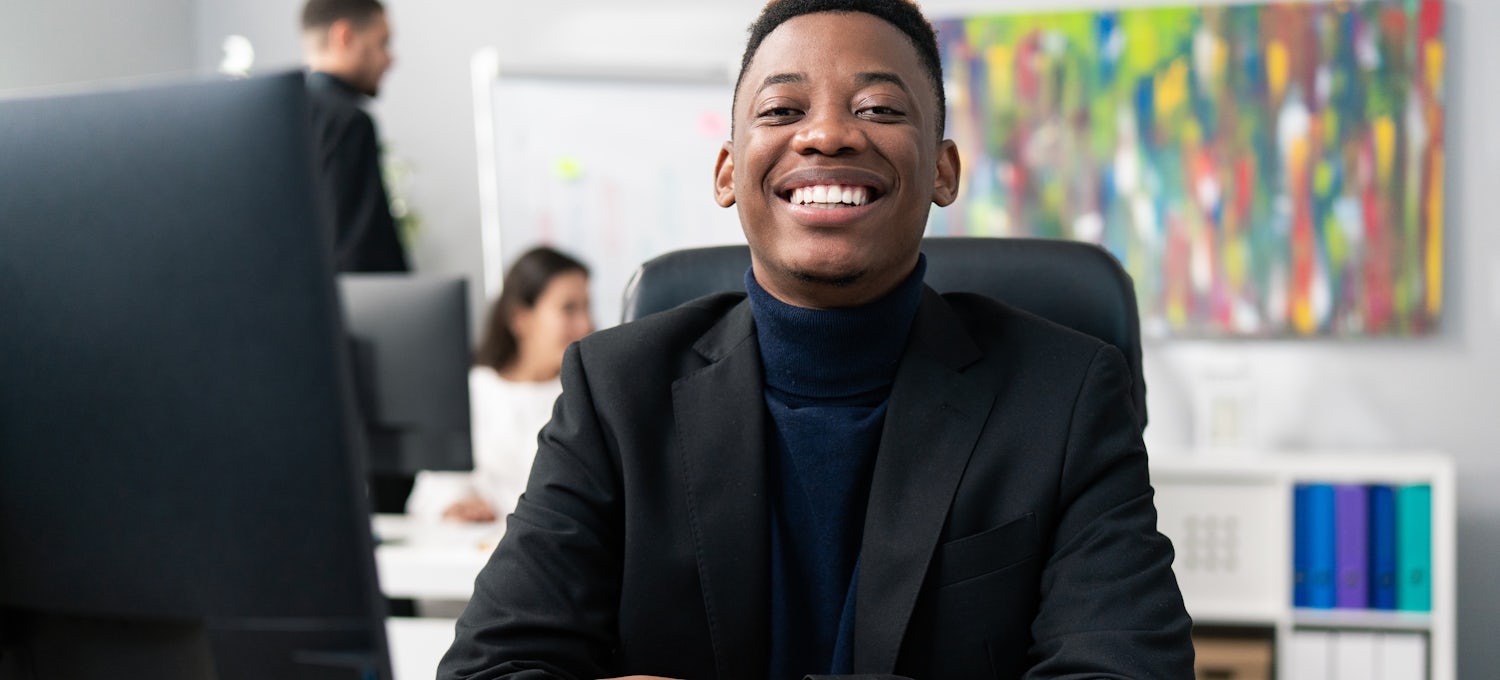 [Featured image] A Bachelor of Business Administration (BBA) student in a black jacket sits in front of a desktop computer and smiles at the camera.