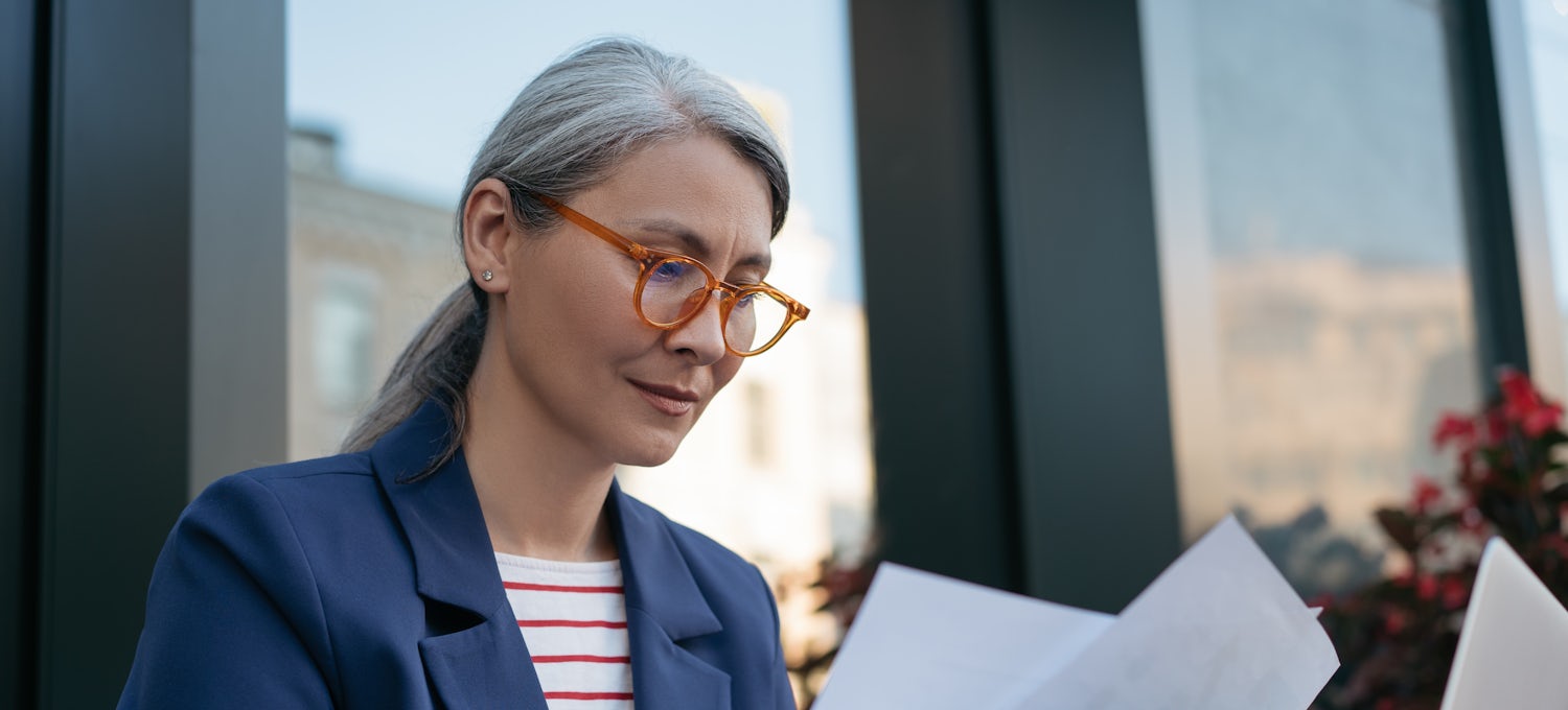 [Featured Image] A person wearing a blue jacket, red and white top, and glasses works on her resume. 