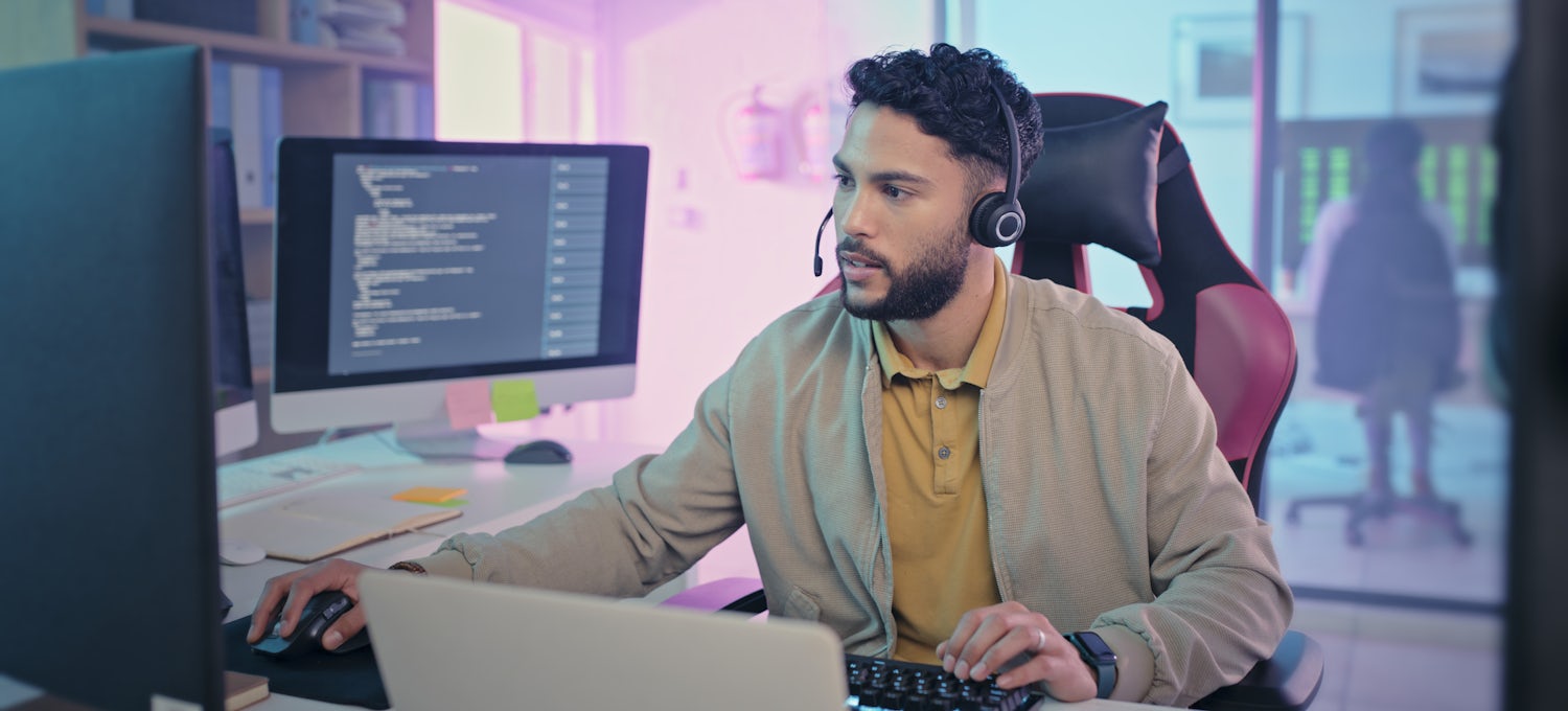 [Featured Image] Man sitting at a desk with several computer monitors in his office, wearing a headset and working intently at his job as a cloud security engineer.