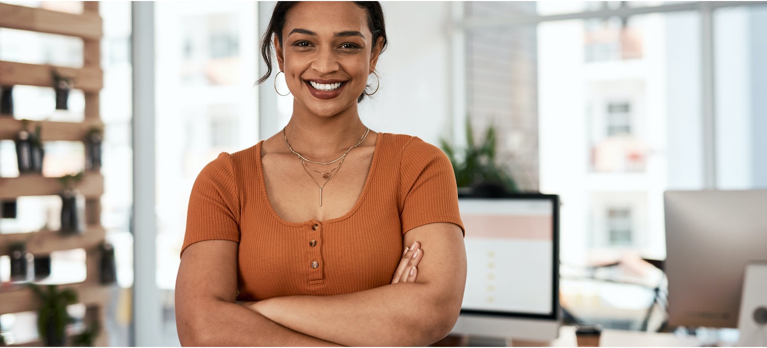 [Imagen destacada] Una sonriente estratega de UX con camisa naranja se cruza de brazos junto a su mesa en una oficina de diseño.