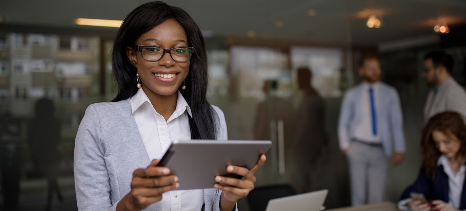 [Featured Image] A woman in business casual clothing is holding a tablet and smiling with three coworkers in the background. 