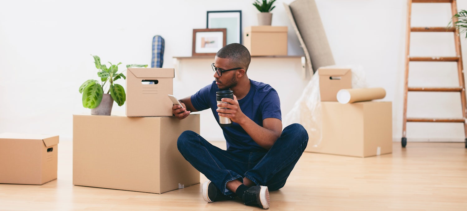 [Featured Image] A man checks his phone in his new home for job openings after moving without a job.