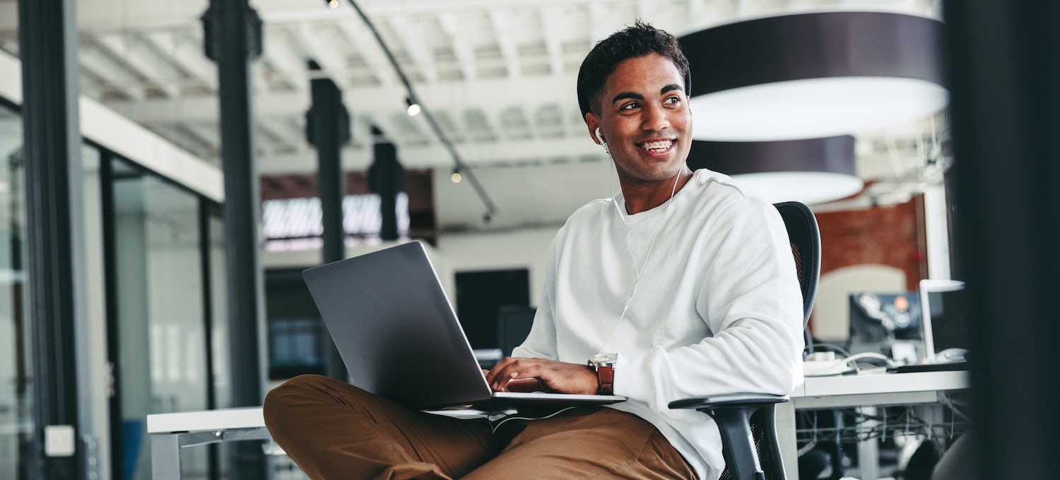 [Featured image] Business degree student sits in a computer lab with a laptop in his lap and is smiling.