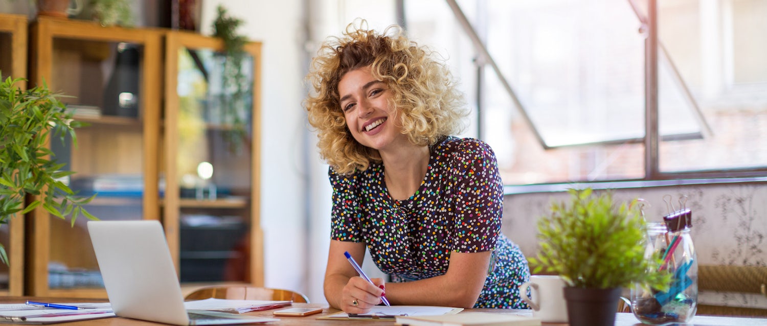 Recent marketing degree graduate, smiling, with curly blonde hair, wearing a dotted work dress, and looking at the camera, leans over a desk with an open laptop, work papers all around, and two plants decorating the space.