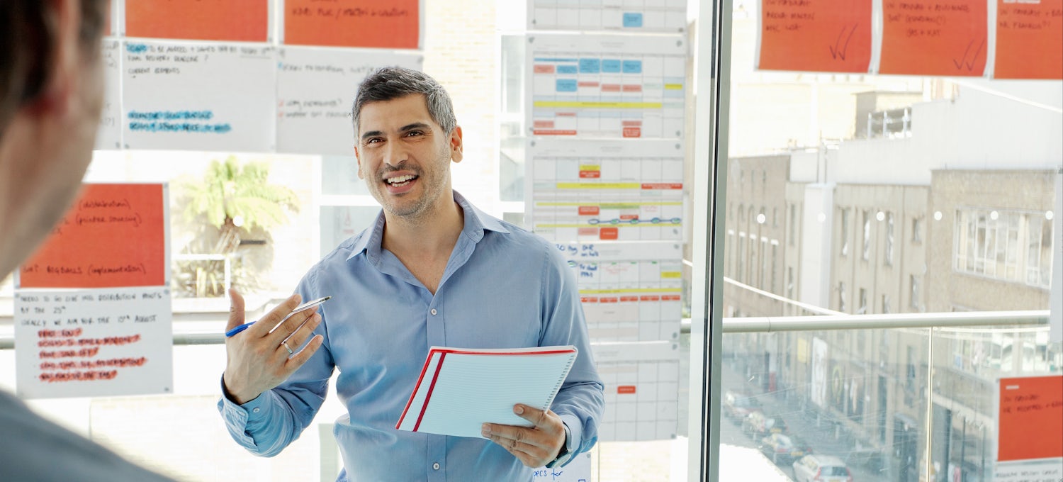 [Featured Image] A project manager stands in front of multicolored charts and discusses Gantt charts with colleagues.