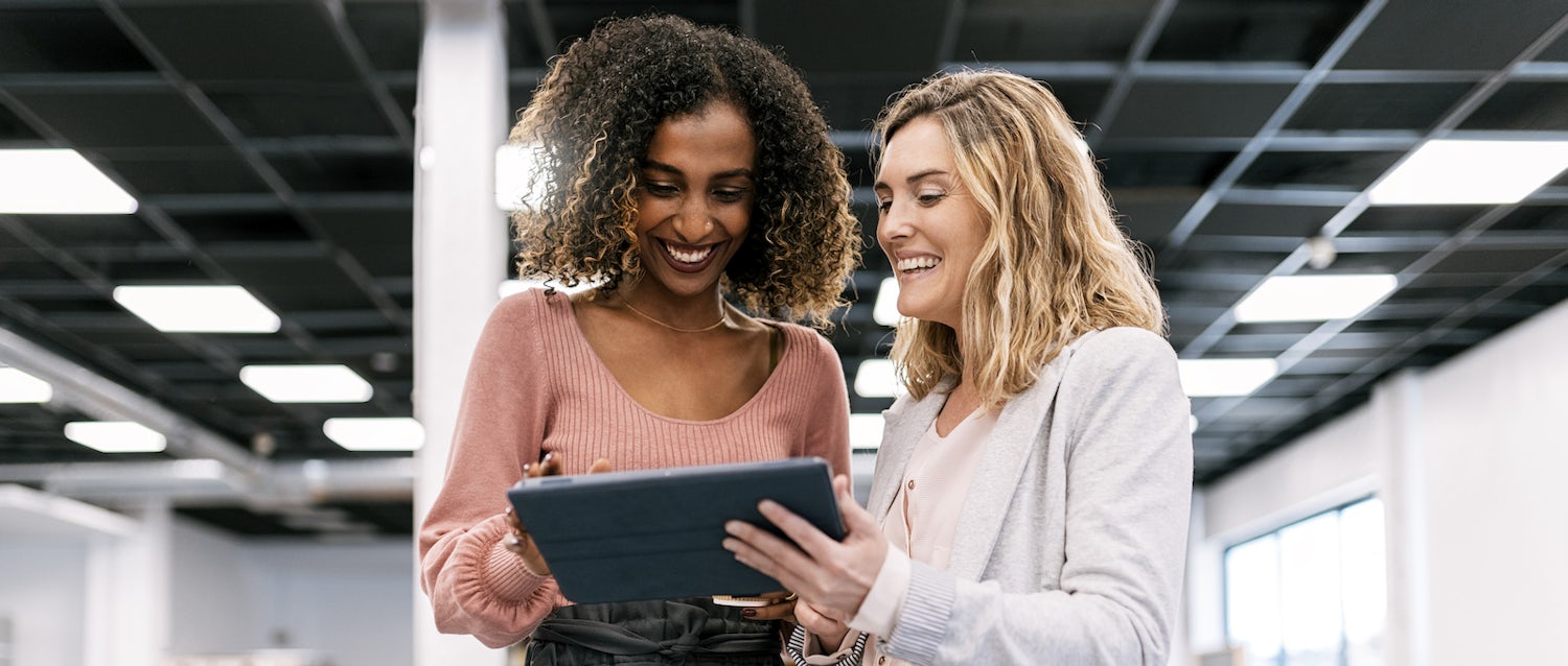 Two women in an office smile while looking at a tablet.