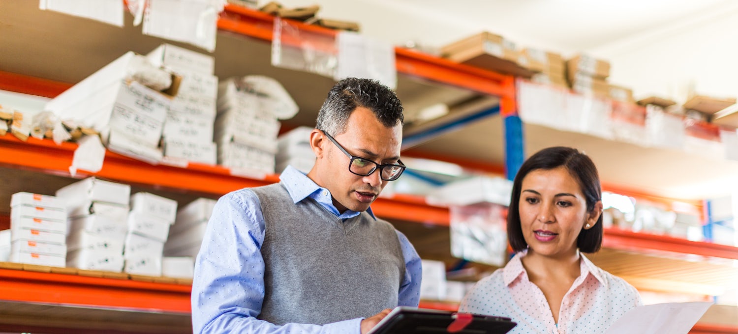 [Featured Image]:  A person in a blue shirt and gray sweater vest stands in a warehouse lined with orange shelving and discusses with someone in a pink shirt whether they need a program manager or a project manager for a job.