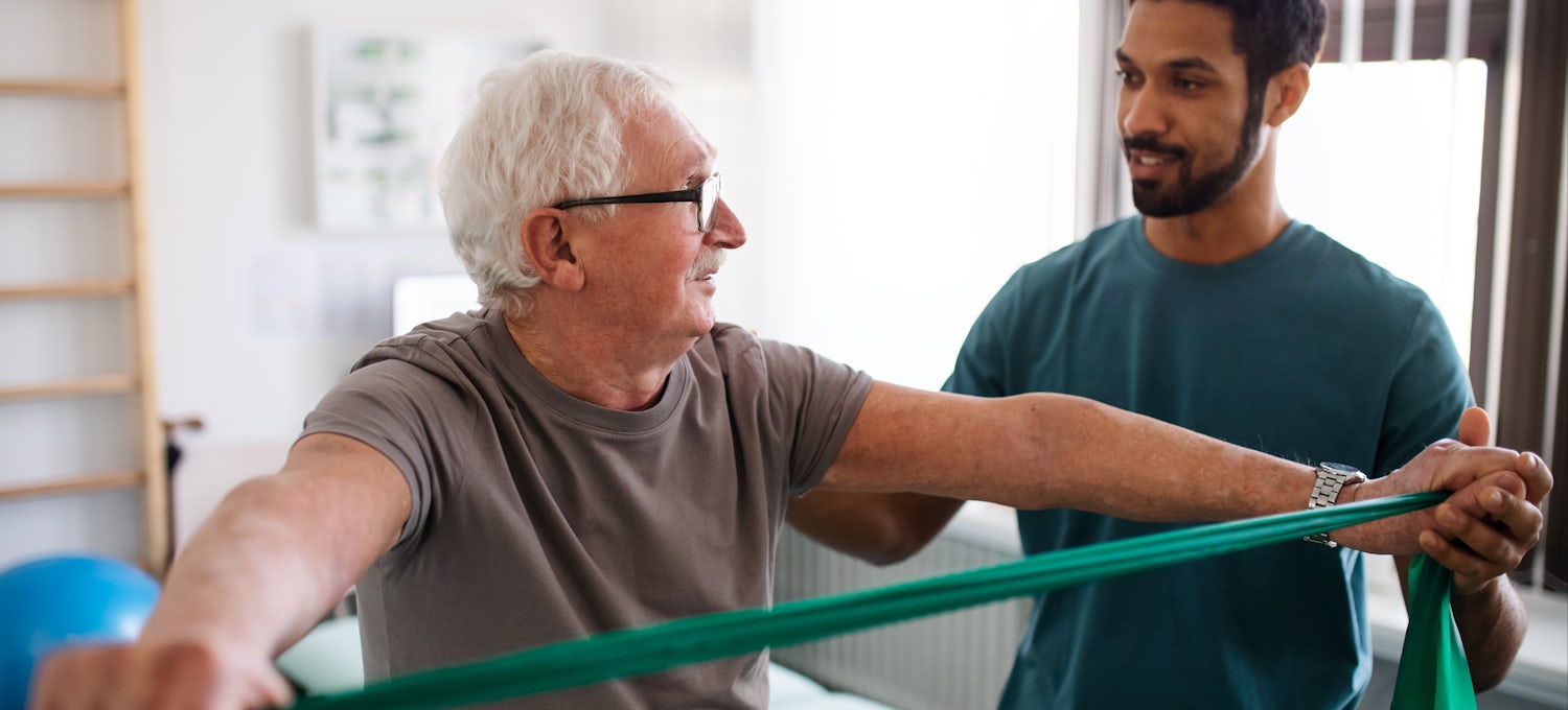 [Featured image] A physical therapist with a degree assists an elderly patient with the help of a resistance band.