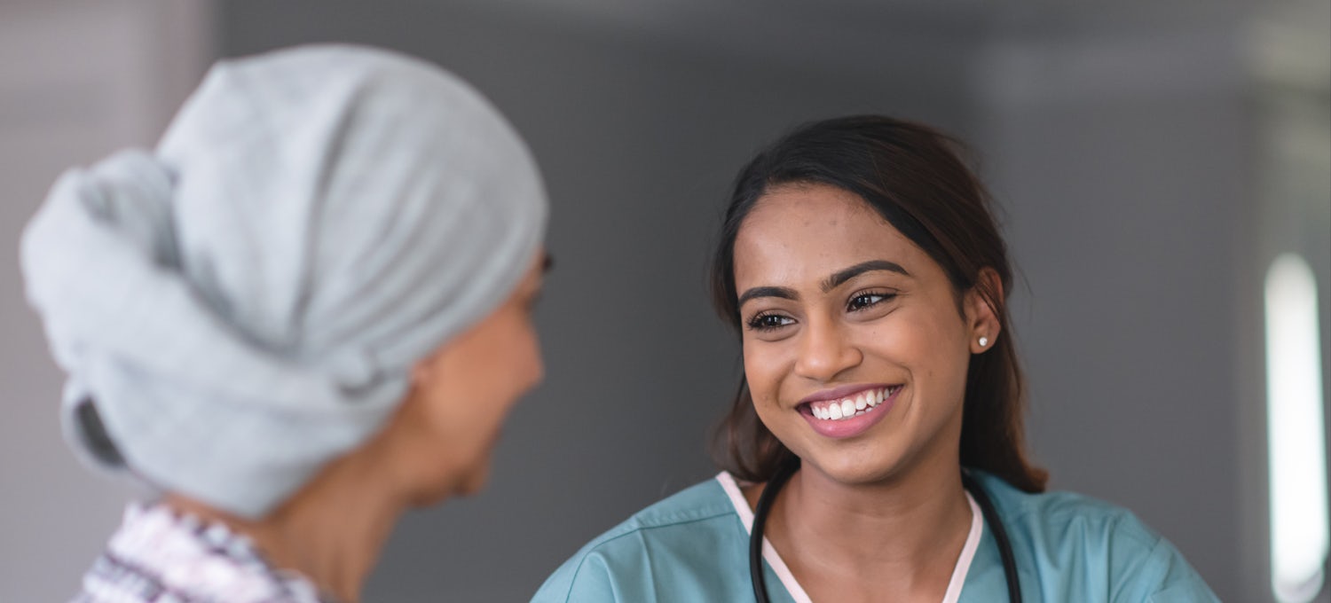 [Featured Image]: A woman with long brown hair and smiling.  She is wearing blue scrubs with white trim. She has a stethoscope around her neck. She is talking to a woman wearing a multi-colored sweater and a head covering.