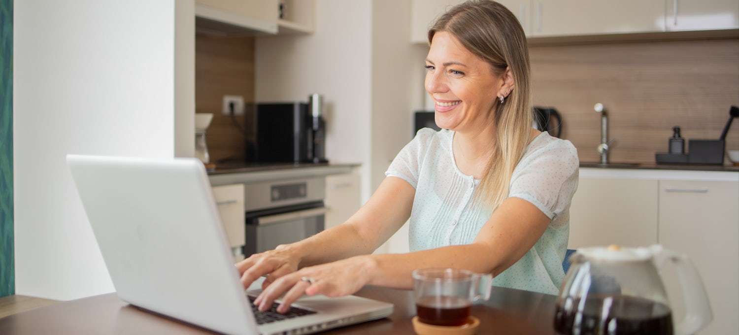 [Featured Image] A woman sits in her kitchen and uses her laptop to research whether she should earn IT certifications or a degree for her career path. 
