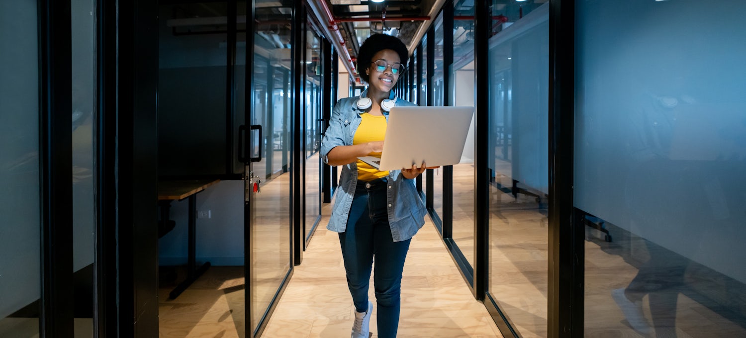 [Featured image] A cloud engineer walks down a hallway with her laptop.