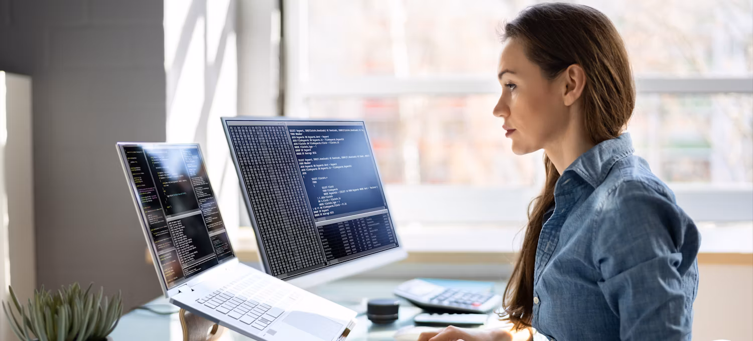 Une personne en chemise bleue est assise à un bureau près d'une fenêtre et travaille sur un code à l'aide d'un ordinateur portable et d'un second écran.