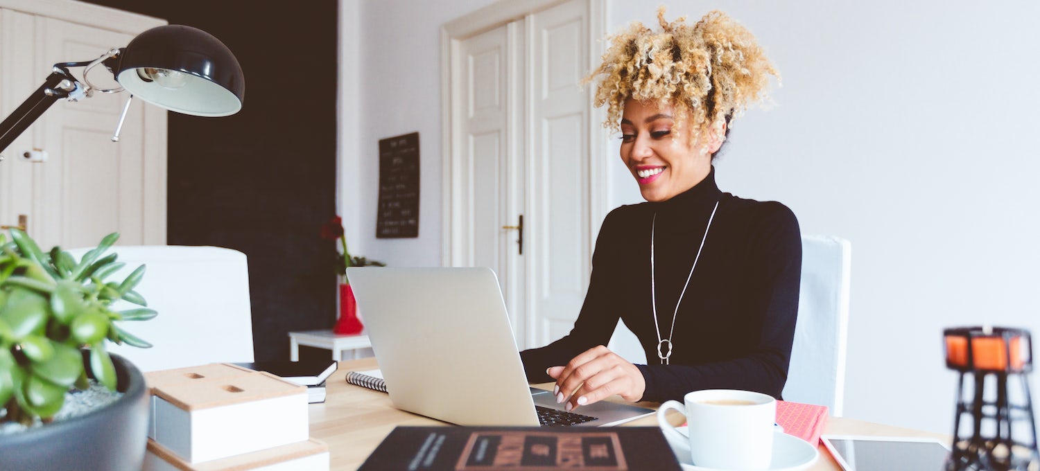 [Imagen destacada] Una mujer sonriente con camisa negra y collar se sienta ante su portátil y trabaja en su portafolio de UX en una oficina muy iluminada.