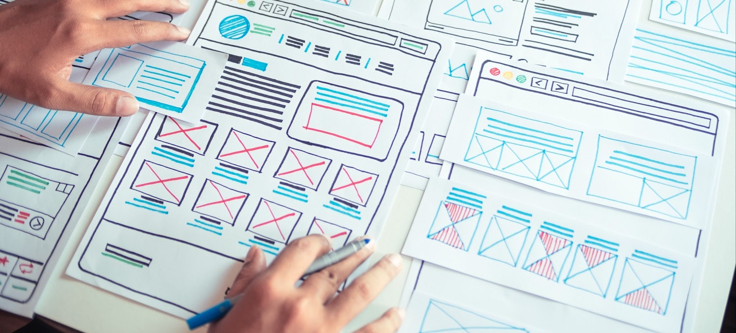 [Featured Image] A UX designer hand draws wireframes with colored markers on sheets of paper at a desk.
