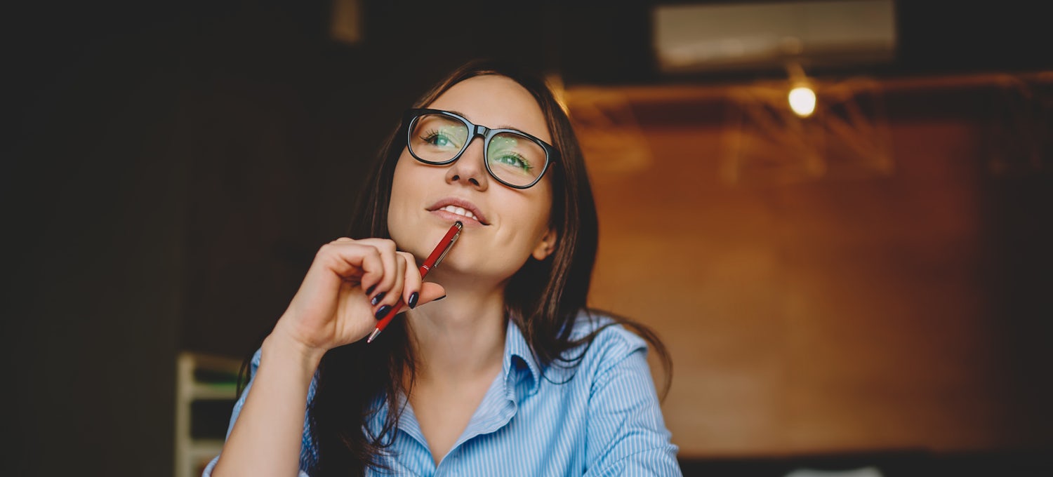 [Featured Image]: A woman with long dark hair wearing glasses and a blue shirt has a pen up to her chin and she is thinking about her 30-60-90 day plan.