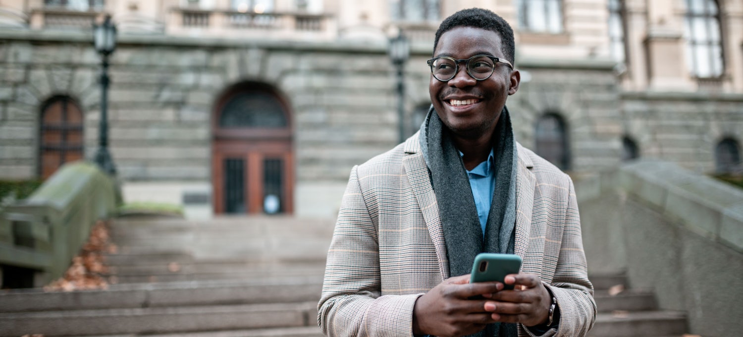 [Featured image] A man in a jacket, scarf and glasses holds a cell phone and smiles looking off-camera.