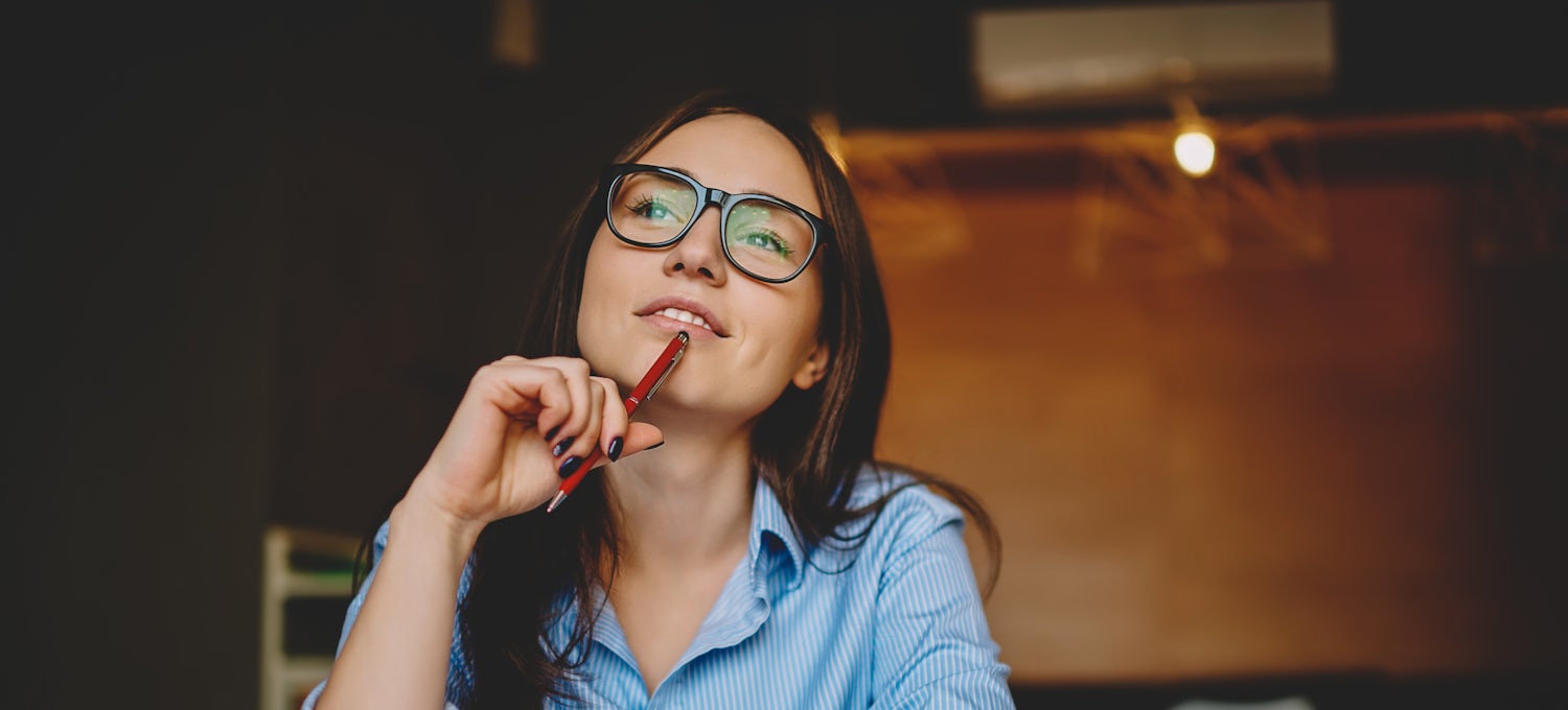[Featured Image]: A woman wearing a blue blouse and glasses, with a pen to chin, thinking about her career path.