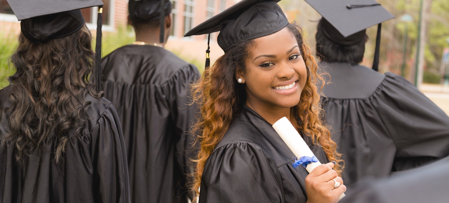 [Featured image] A woman in her cap and gown holds her diploma and smiles after getting her bachelor’s degree with her classmates.