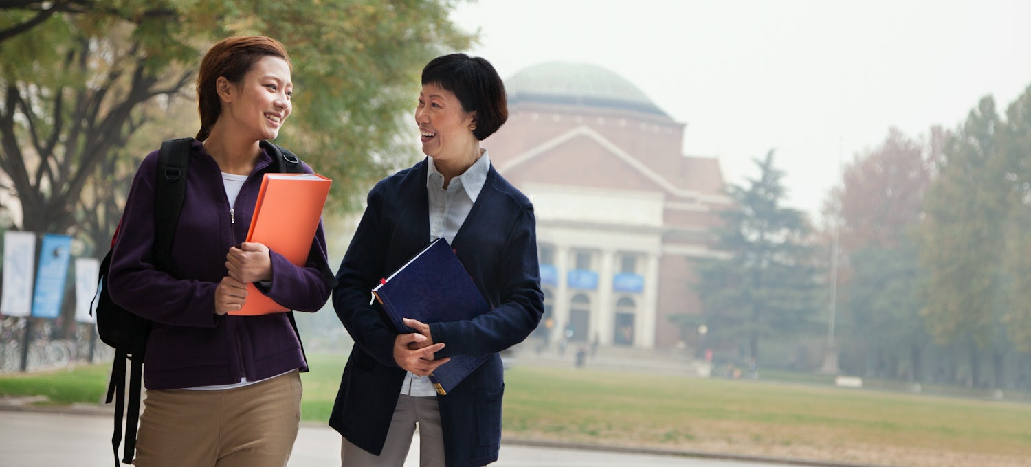 [Featured image] A marketing degree student with a backpack and an orange folder walks through a college campus with a female professor wearing a jacket.