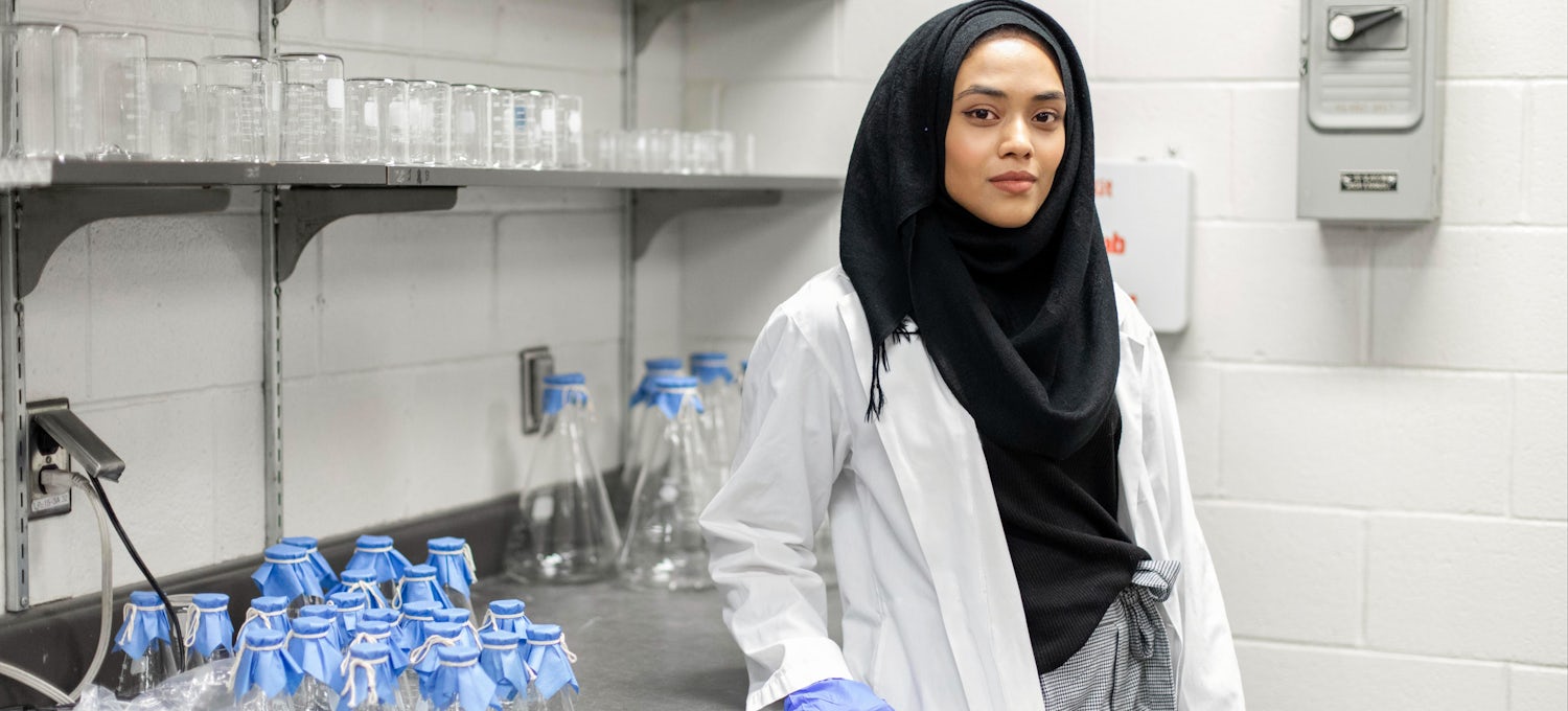 [Featured image] A PharmD student stands in a lab wearing a black headscarf, white lab coat, and blue medical gloves.