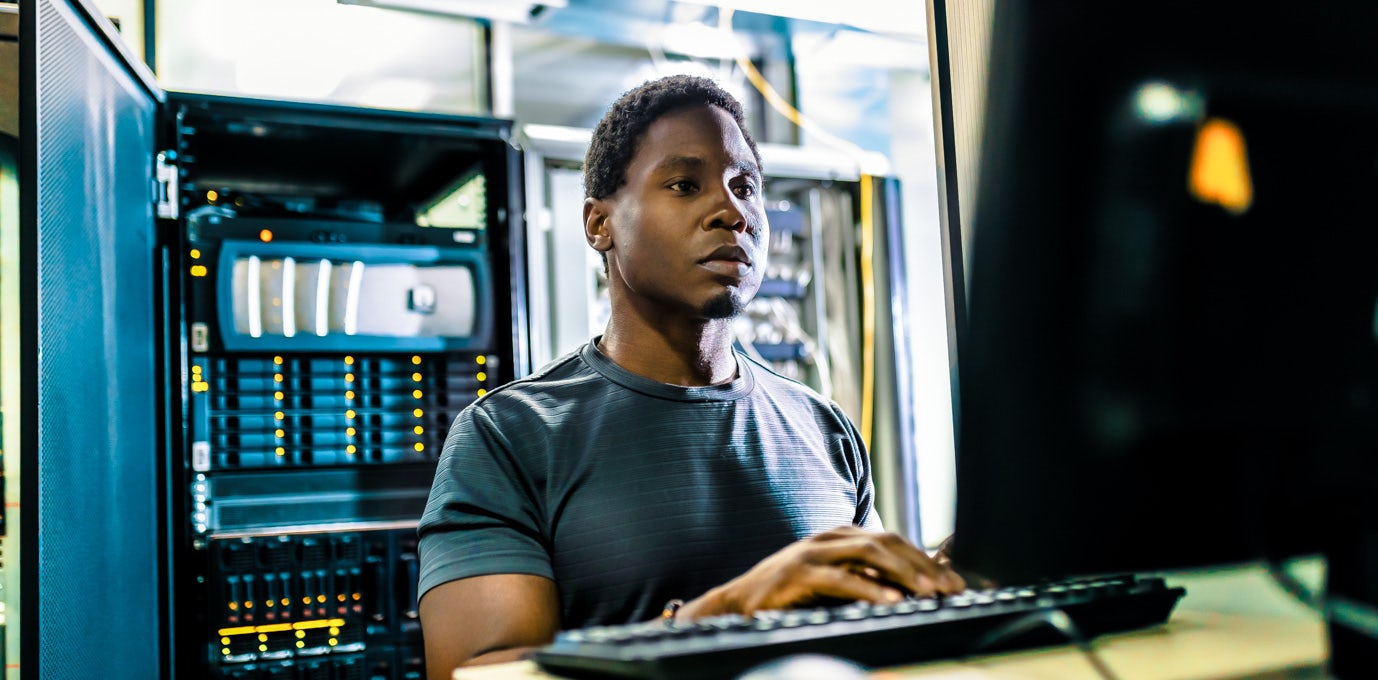 [Featured image] A CompTIA A+-certified IT worker sits in a server room and works on a black desktop computer.
