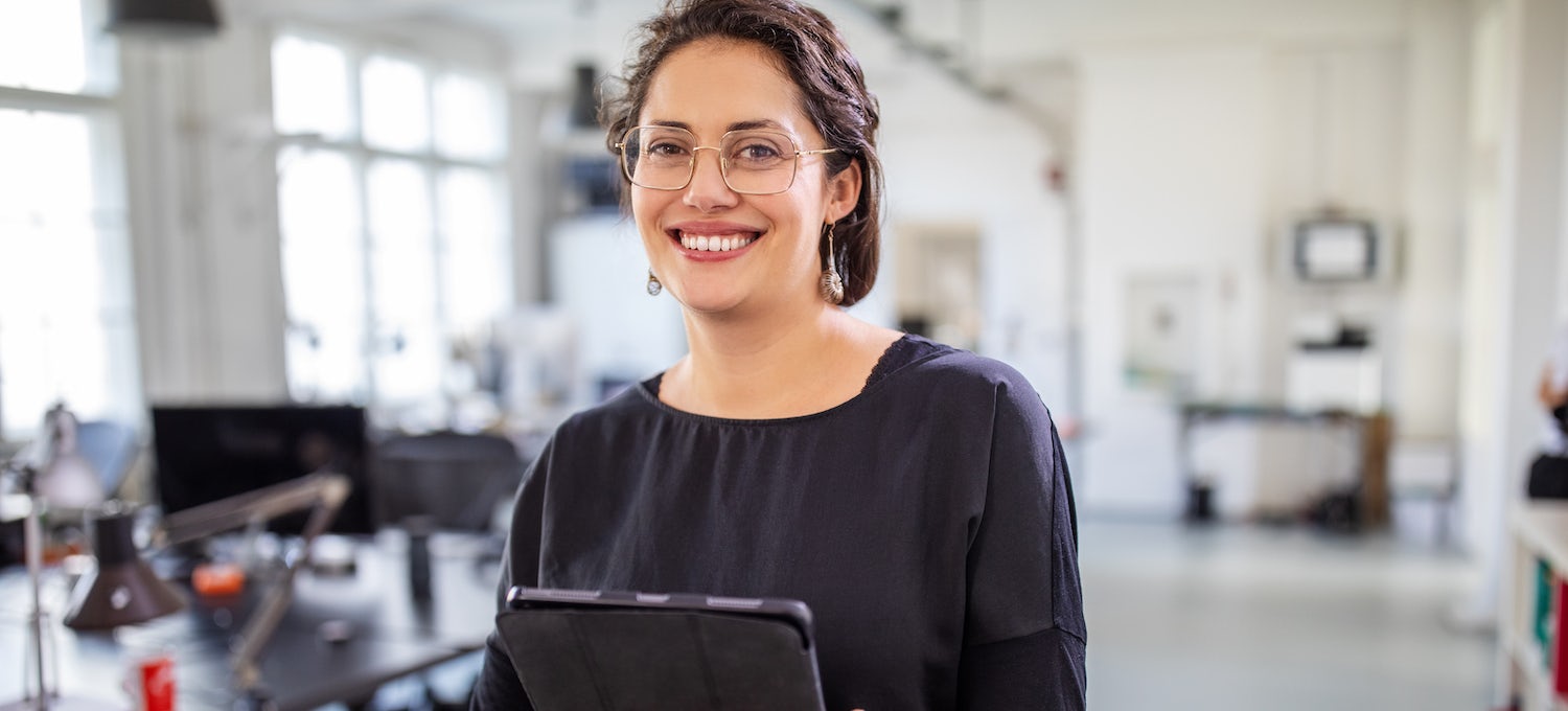 [Featured image] A young woman wearing glasses and holding an iPad stands smiling in an empty office. 
