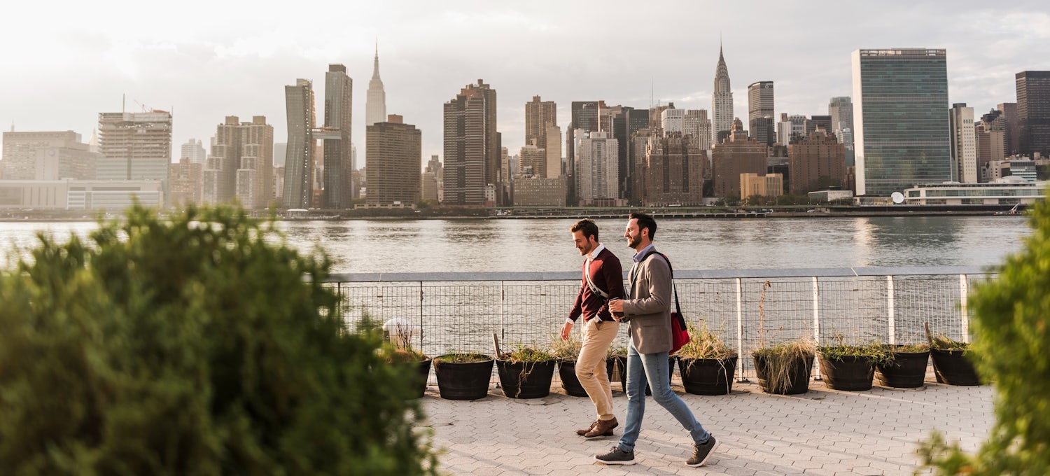 [Featured image] Two mean in business attire walk along the waterfront with the New York City skyline in the background.