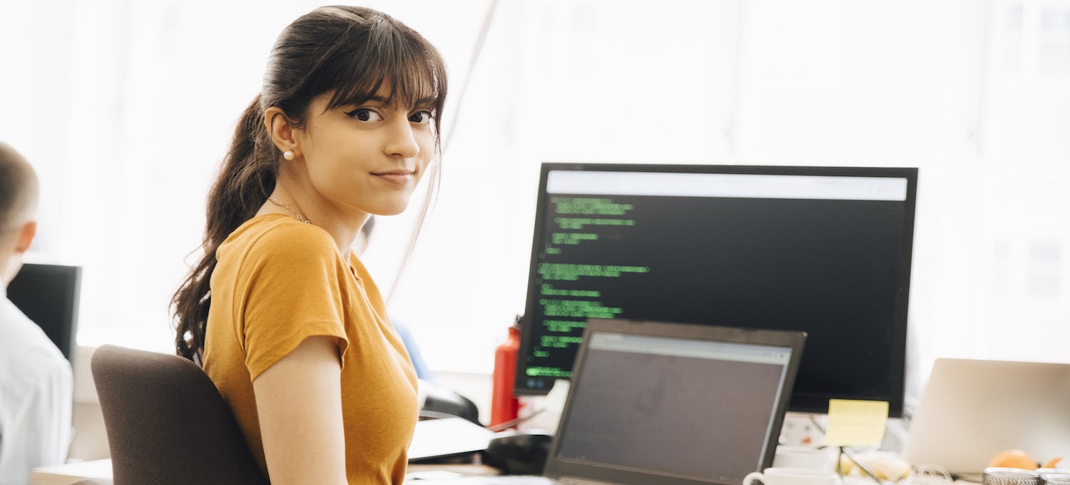 [Featured image] A young woman with her hair in a ponytail and wearing a golden yellow shirt sits in front of a laptop and desktop displaying code. 
