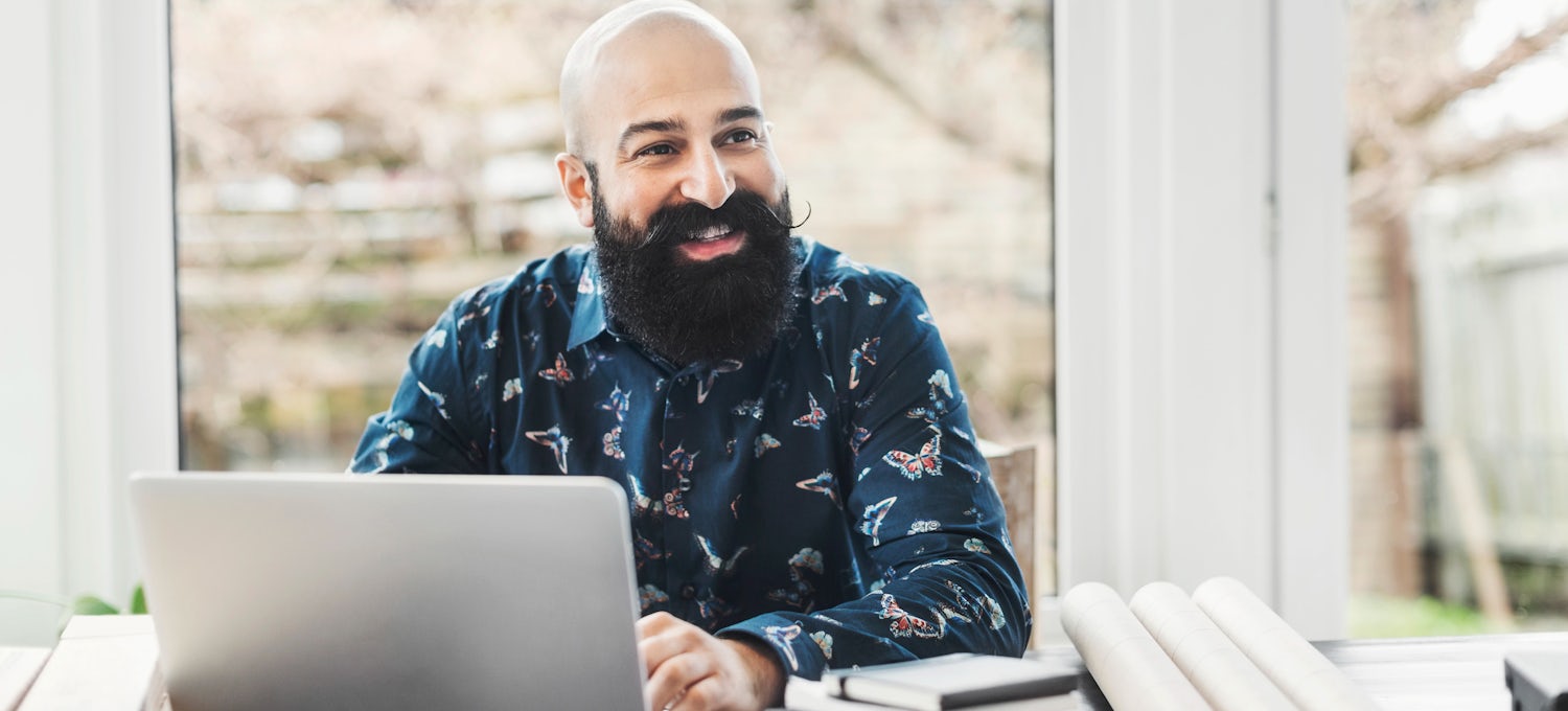 [Featured Image] A person with a beard sits at a desk in front of a laptop smiling. 
