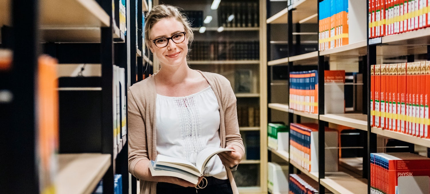[Featured Image] An English major stands in a library learning against a bookshelf while holding two books.