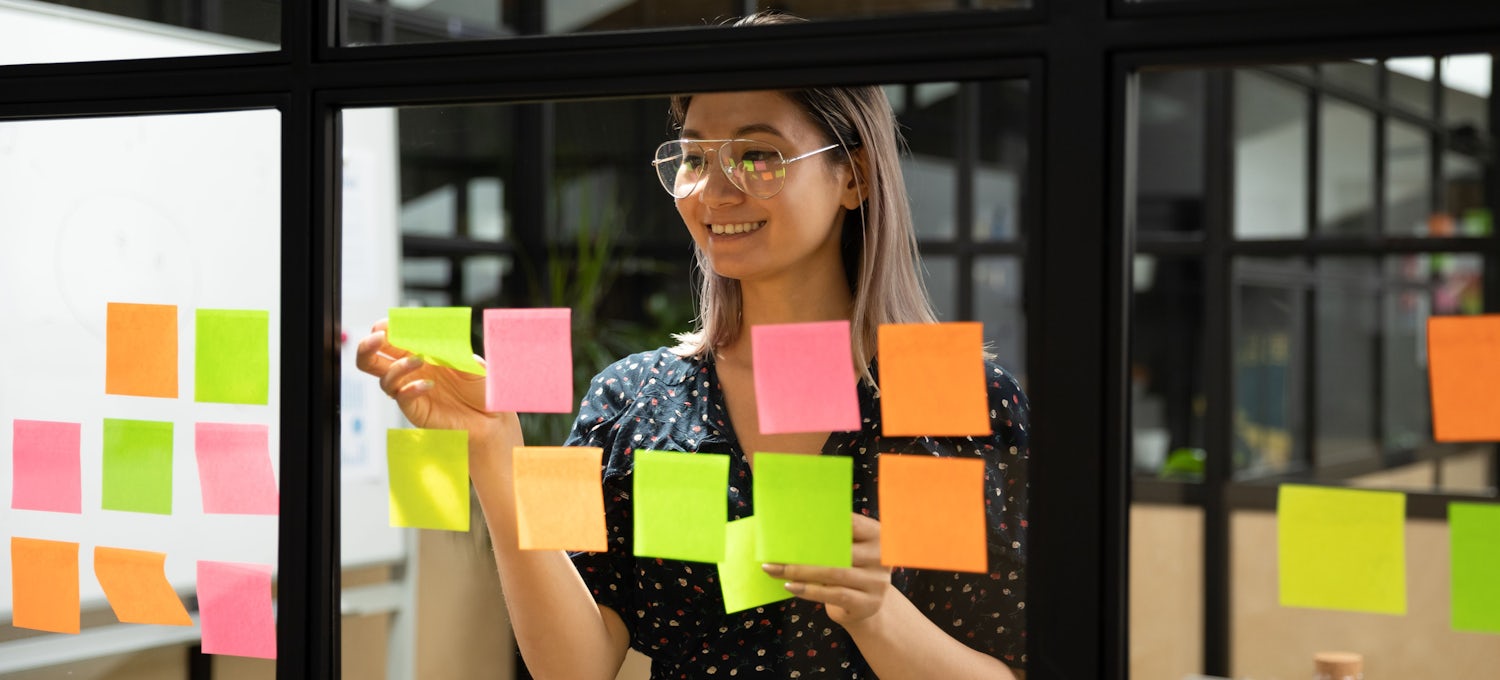 [Featured Image] A Scrum Master smiles and puts post-it notes on a window.  