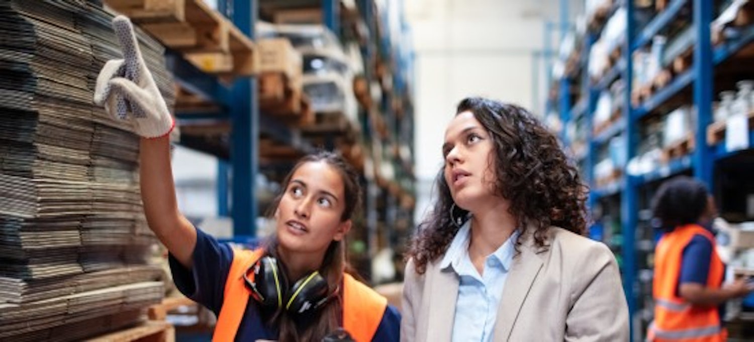 [Featured image] Two product managers are wearing an orange vest, and gloves looking at warehouse products. 